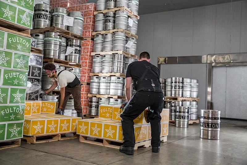 Workers using Ergodyne back supports while lifting heavy boxes in a warehouse – Pryme Australia ergonomic safety.