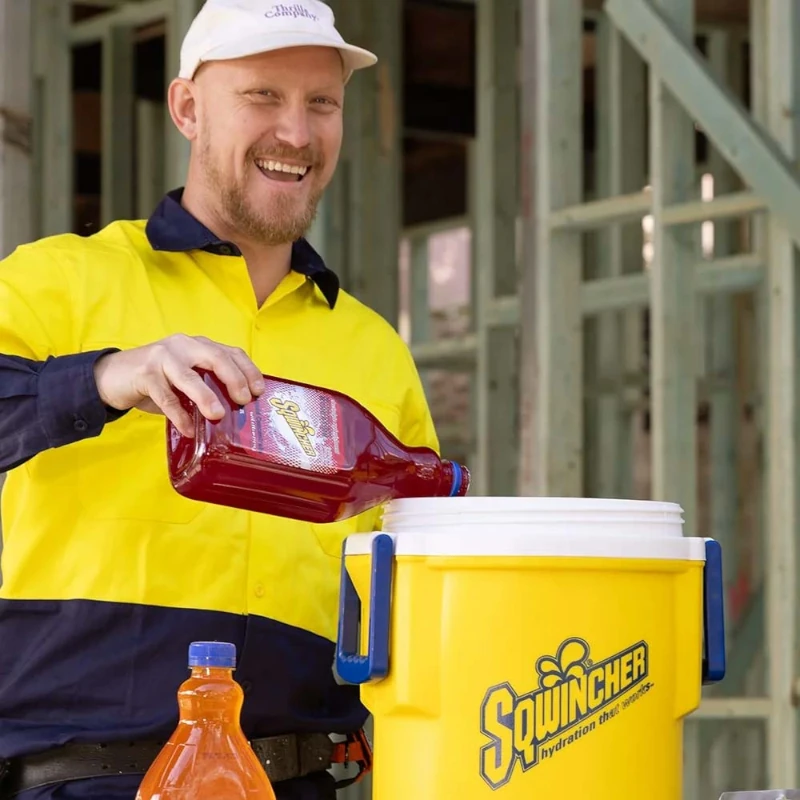 Worker pouring Sqwincher concentrate into large yellow cooler for team hydration on construction site.
