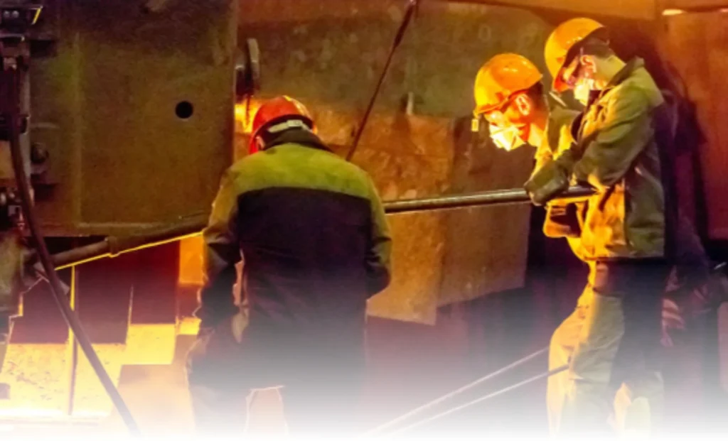 Industrial workers wearing protective helmets and gloves working near molten metal in extreme heat.